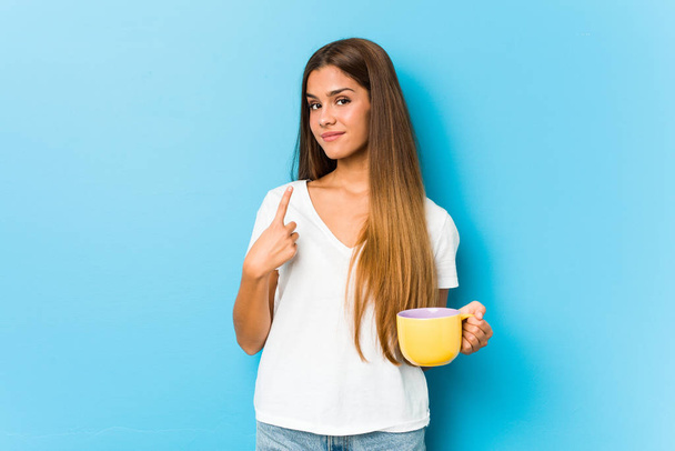 Jeune femme caucasienne tenant une tasse de café pointant du doigt vers vous comme si vous invitant se rapprocher
. - Photo, image