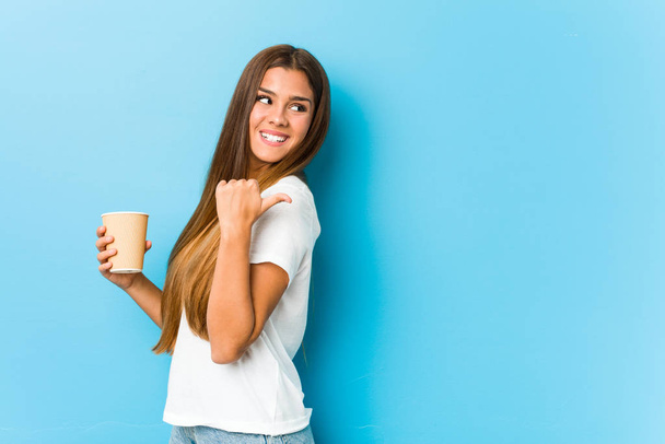 junge hübsche kaukasische Frau hält einen Kaffee zum Mitnehmen zeigt mit dem Daumen weg, lacht und unbeschwert. - Foto, Bild