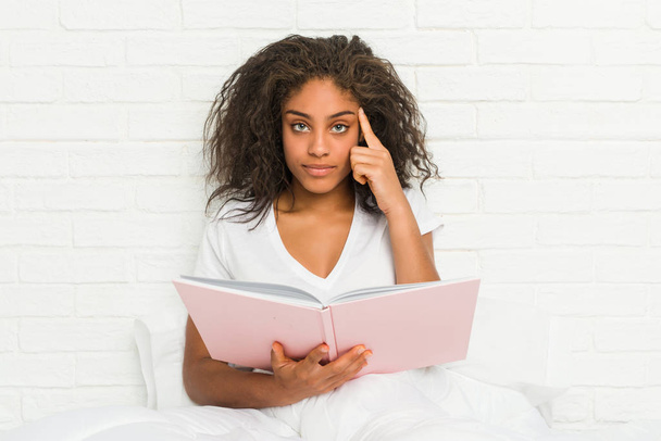 Joven mujer afroamericana sentada en la cama estudiando señalando su sien con el dedo, pensando, enfocada en una tarea
. - Foto, Imagen