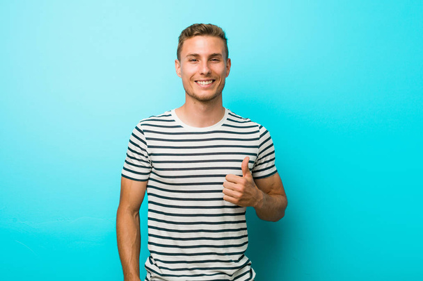 Nuori valkoihoinen mies vastaan sininen seinä hymyilee ja nostaa peukalo ylös
 - Valokuva, kuva
