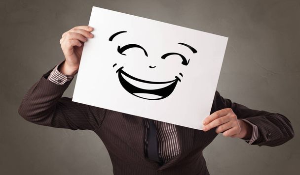 Persona sosteniendo un papel delante de su cara con emoticono garabato
 - Foto, Imagen