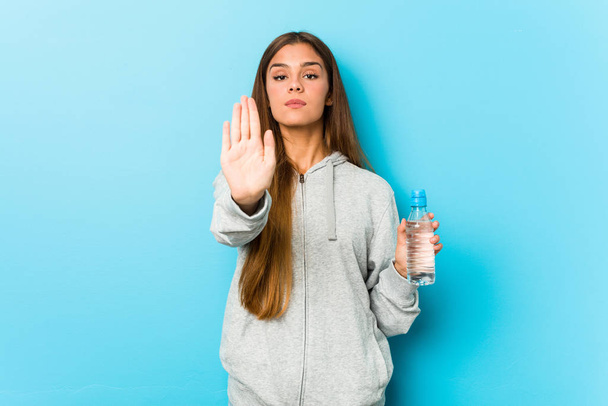 Mulher fitness jovem segurando uma garrafa de água de pé com a mão estendida mostrando sinal de parada, impedindo-o
. - Foto, Imagem