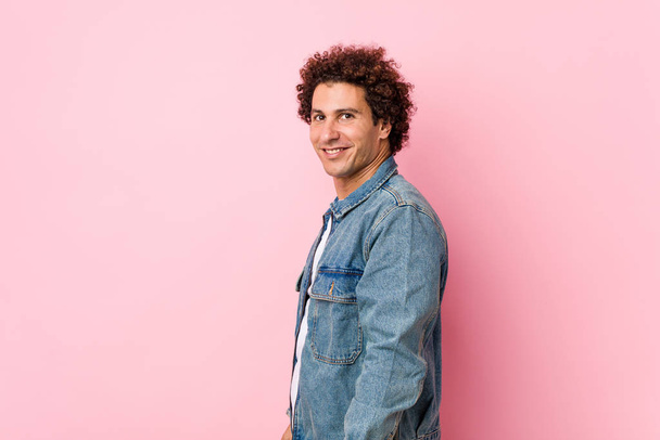 Curly homme mature portant une veste en denim sur fond rose regarde de côté souriant, gai et agréable
. - Photo, image