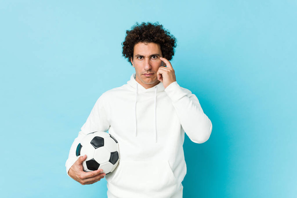 Uomo sportivo in possesso di un pallone da calcio che punta il suo tempio con il dito, pensando, focalizzato su un compito
. - Foto, immagini