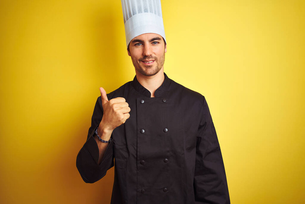 Young chef man wearing uniform and hat standing over isolated yellow background doing happy thumbs up gesture with hand. Approving expression looking at the camera showing success. - Fotó, kép