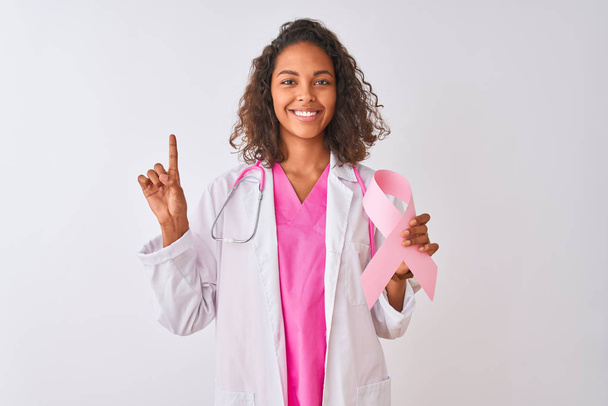 junge brasilianische Ärztin hält Krebsband über isoliertem weißem Hintergrund und überrascht mit einer Idee oder Frage, die mit dem Zeigefinger auf glückliches Gesicht zeigt, Nummer eins - Foto, Bild