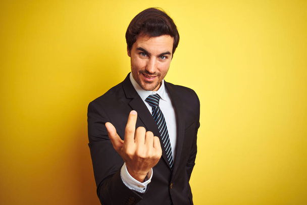 Young handsome businessman wearing suit and tie standing over isolated yellow background Beckoning come here gesture with hand inviting welcoming happy and smiling - Photo, Image
