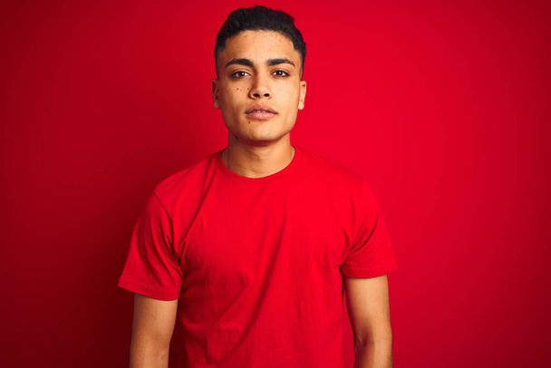 Молодий бразіліан, одягнений в футболку, стоїть над ізольованим червоним фоном Розслаблений з серйозним виразом обличчя. Простий і природний погляд на камеру.. - Фото, зображення