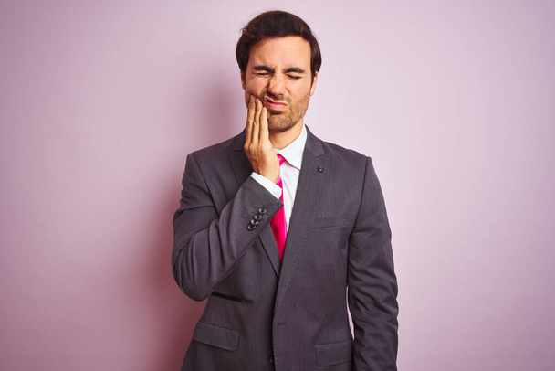 Young handsome businessman wearing suit and tie standing over isolated pink background touching mouth with hand with painful expression because of toothache or dental illness on teeth. Dentist concept. - 写真・画像