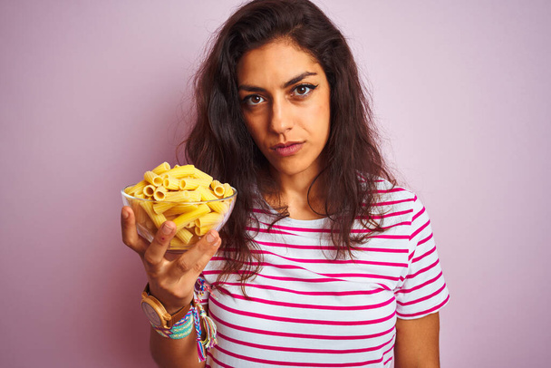 Young beautiful woman holding bowl with dry macaroni pasta over isolated pink background with a confident expression on smart face thinking serious - Photo, Image