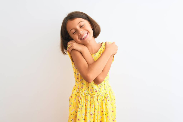 Mladý krásný dítě dívka na sobě žluté květinové šaty stojící nad izolovaným bílým pozadím objímající se šťastný a pozitivní, usmívající se sebevědomě. Sebeláska a sebepéče - Fotografie, Obrázek