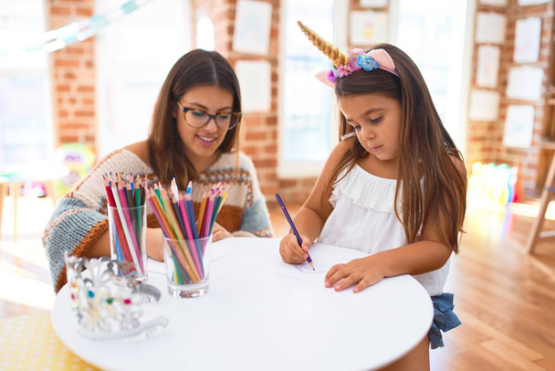 Belle enseignante et tout-petit portant un diadème licorne en utilisant du papier et des crayons autour de beaucoup de jouets à la maternelle
 - Photo, image