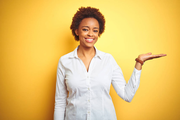 African american business woman over isolated yellow background smiling cheerful presenting and pointing with palm of hand looking at the camera. - Zdjęcie, obraz
