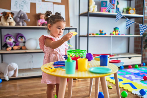 Молодой красивый ребенок играет со столовыми приборами и пищевыми игрушками на столе в детском саду
 - Фото, изображение