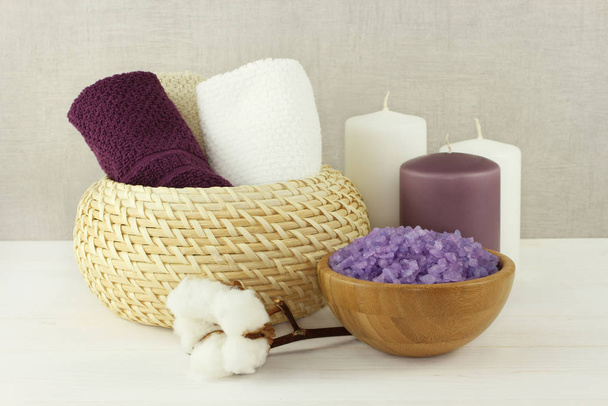 Lavender sea salt for bath in a bamboo bowl, cotton towels in a wicker vase and candles stand on a white wooden table against a gray wall - Photo, Image
