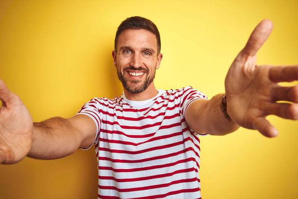 Mladý pohledný muž v neformálním červeném pruhovaném tričku na žlutém izolovaném pozadí, dívající se na kameru, usmívající se s otevřenou náručí k obejmutí. Veselý výraz objímající štěstí. - Fotografie, Obrázek