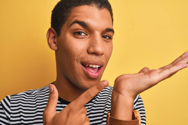 Joven hombre árabe guapo con camiseta a rayas azul marino sobre fondo amarillo aislado muy feliz señalando con la mano y el dedo
 - Foto, Imagen