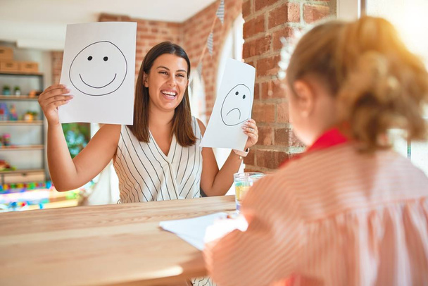 Hermosa psicóloga y niña rubia vistiendo uniforme escolar sentada en el escritorio haciendo terapia usando emociones emoji en la sala de consulta
 - Foto, Imagen