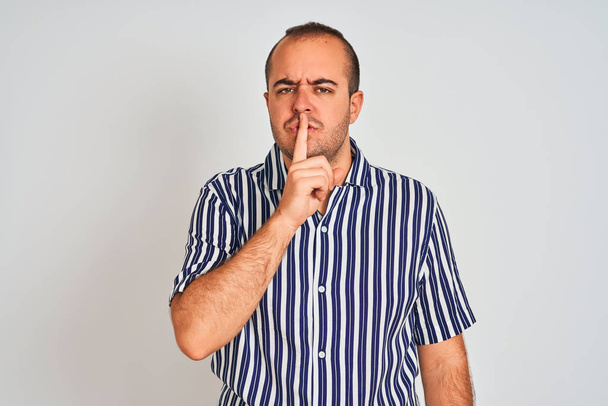 Νεαρός άνδρας με μπλε ριγέ πουκάμισο στέκεται πάνω από απομονωμένο λευκό φόντο ζητώντας να είναι ήσυχο με το δάχτυλο στα χείλη. Σιωπή και μυστική έννοια. - Φωτογραφία, εικόνα