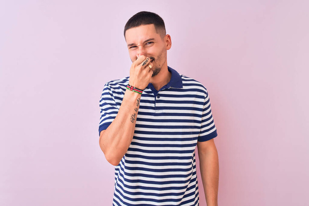 Νεαρός όμορφος άντρας που φοράει ναυτικό ριγέ μπλουζάκι πάνω από ροζ απομονωμένο φόντο μυρίζοντας κάτι βρωμερό και αηδιαστικό, ανυπόφορη μυρωδιά, κρατώντας ανάσα με δάχτυλα στη μύτη. Έννοια κακών οσμών. - Φωτογραφία, εικόνα