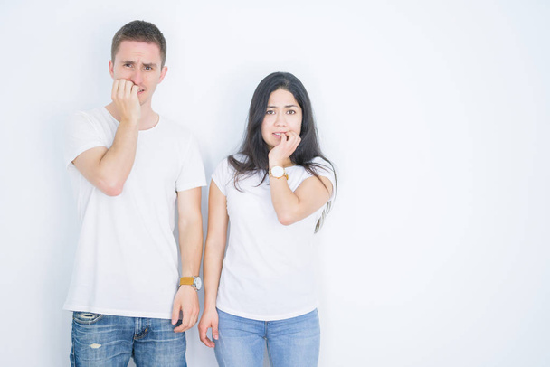 Молода красива пара в повсякденній футболці стоїть над ізольованим білим тлом, виглядає напруженим і нервує руками на роті, кусаючи цвяхи. Проблема тривоги
. - Фото, зображення