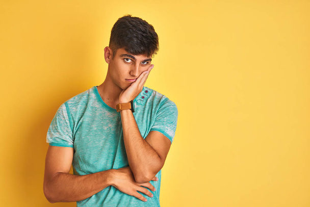 Jovem indiano vestindo camiseta verde em pé sobre fundo amarelo isolado pensando cansado e entediado com problemas de depressão com os braços cruzados
. - Foto, Imagem