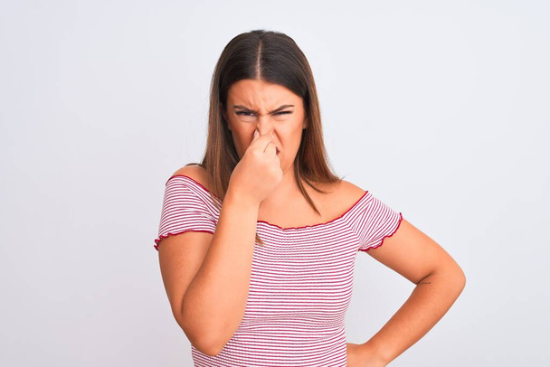 Portrait of beautiful young woman standing over isolated white background smelling something stinky and disgusting, intolerable smell, holding breath with fingers on nose. Bad smells concept. - Photo, image