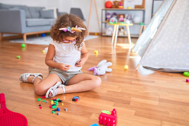 Beautiful toddler wearing glasses and unicorn diadem sitting on the floor playing with building blocks at kindergarten - Фото, изображение