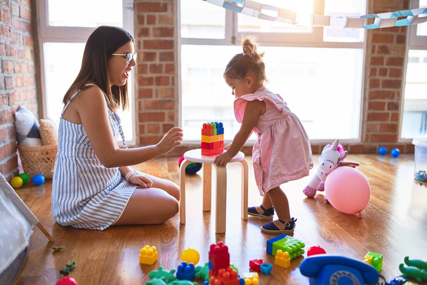 Joven hermosa maestra y niño pequeño jugando con bloques de construcción de juguete en la mesa en el jardín de infantes
 - Foto, imagen