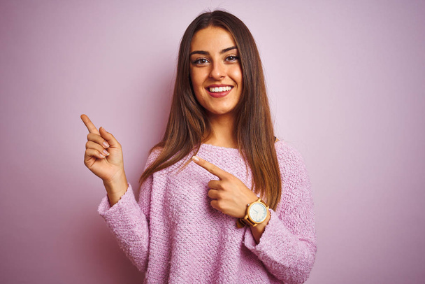 Молода красива жінка в повсякденному светрі стоїть над ізольованим рожевим тлом, посміхаючись і дивлячись на камеру, спрямовану двома руками і пальцями в сторону
. - Фото, зображення