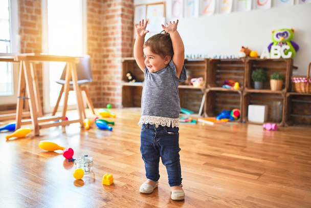 Красивий малюк стоїть з піднятими руками усміхнений навколо багатьох іграшок в дитячому саду
 - Фото, зображення