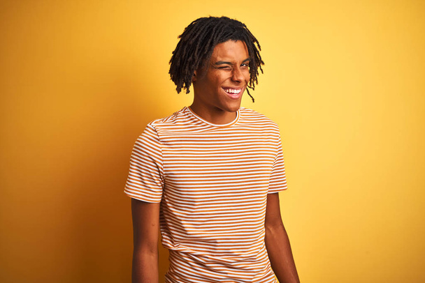 Afro człowiek z dredami w paski koszulka stoi nad odizolowanym żółtym tle mruga patrząc w aparat z seksowną ekspresją, wesoły i szczęśliwy twarz. - Zdjęcie, obraz