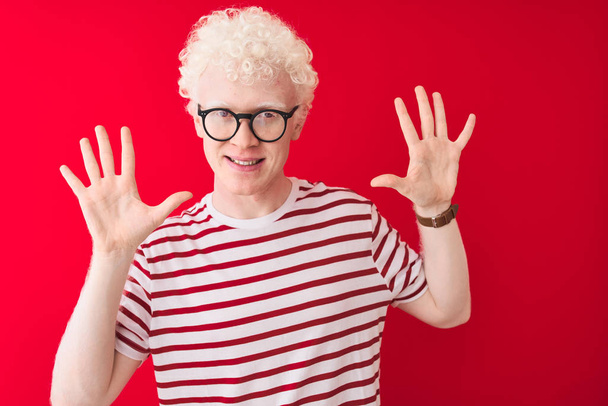 Молодий альбіно блондин в смугастій футболці і окулярах над ізольованим червоним тлом, показуючи і вказуючи пальцями номер десять, посміхаючись впевнено і щасливо
. - Фото, зображення