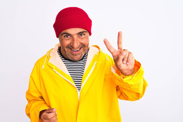 Mężczyzna w średnim wieku ubrany w płaszcz przeciwdeszczowy i wełniany kapelusz stojący nad odizolowanym białym tłem pokazujący i wskazujący palcami numer dwa uśmiechnięty pewny siebie i szczęśliwy. - Zdjęcie, obraz