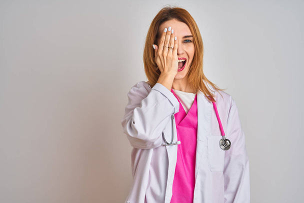 Руда біла жінка-лікар в рожевому стетоскопі над ізольованим тлом, що покриває одне око рукою, впевнена посмішка на обличчі та несподівані емоції
. - Фото, зображення