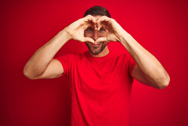 Молодой красивый мужчина в повседневной футболке на красном изолированном фоне делает форму сердца с рукой и пальцами, улыбаясь, глядя сквозь знак
 - Фото, изображение