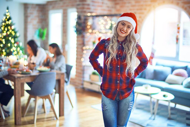 Молодая красивая женщина улыбается счастливой и уверенной. Стоя с улыбкой на лице в сант шляпе празднуя Рождество с семьей дома
 - Фото, изображение