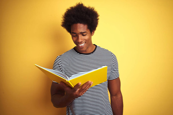 Afro-amerikanischer Student liest Buch vor isoliertem gelben Hintergrund mit einem glücklichen Gesicht stehend und lächelnd mit einem selbstbewussten Lächeln, das Zähne zeigt - Foto, Bild