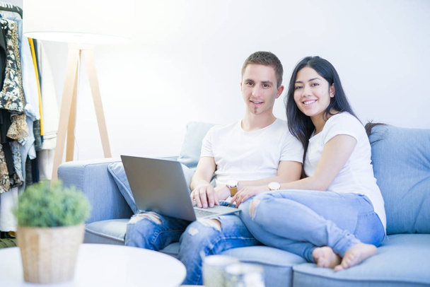 junges schönes Paar sitzt mit Laptop auf dem Sofa im neuen Zuhause um Kartons herum - Foto, Bild