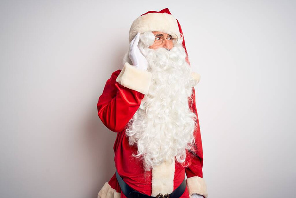 Gut aussehender Mann mittleren Alters im Weihnachtsmannkostüm, der vor isoliertem weißen Hintergrund steht und lächelnd mit der Hand über dem Ohr Gerüchten oder Klatsch lauscht. Konzept für Gehörlosigkeit. - Foto, Bild