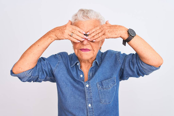 Starší šedovlasá žena v džínové košili a brýlích na izolovaném bílém pozadí zakrývajícím oči s veselýma a legračníma rukama. Slepý koncept. - Fotografie, Obrázek