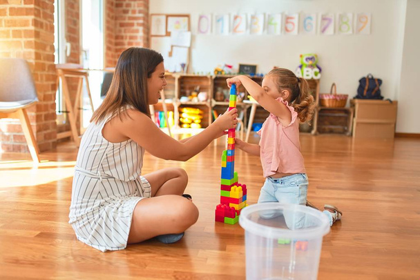 Bela professora e loira criança torre de construção menina usando blocos de plástico no jardim de infância
 - Foto, Imagem