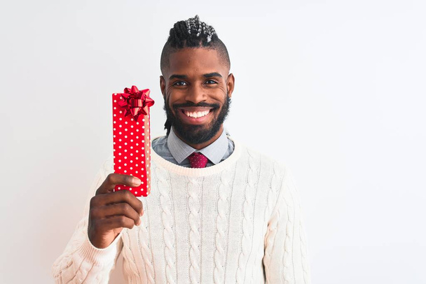 Afrykański Amerykanin z warkoczami trzymający prezent świąteczny na odizolowanym białym tle ze szczęśliwą twarzą stojącą i uśmiechniętą z pewnym uśmiechem pokazującym zęby - Zdjęcie, obraz
