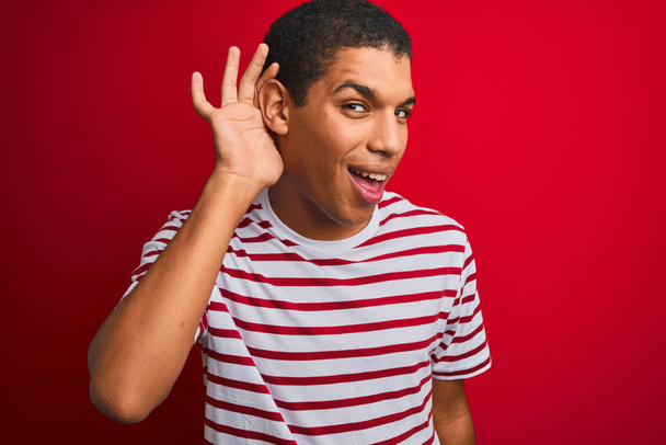 Joven hombre árabe guapo con camiseta a rayas sobre fondo rojo aislado sonriendo con la mano sobre la oreja escuchando un rumor o chismes. Concepto de sordera
. - Foto, Imagen