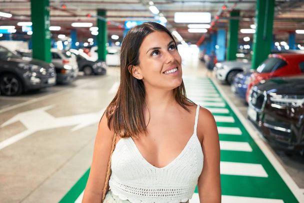 Jovem mulher sorrindo confiante no estacionamento subterrâneo em torno de carros e luzes
 - Foto, Imagem