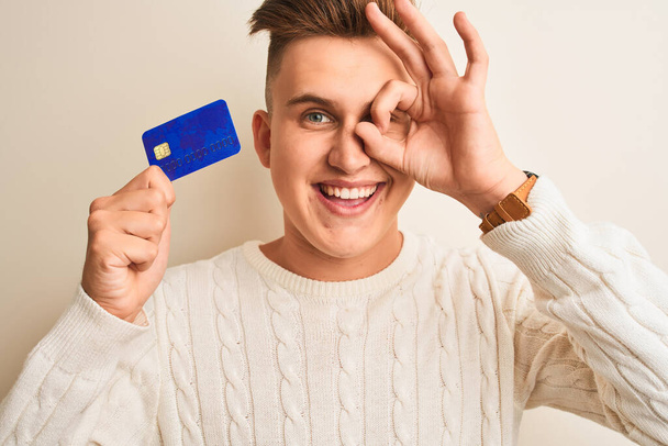 Genç ve yakışıklı bir adam kredi kartını izole edilmiş beyaz arka planda tutarken gülümseyen yüzüyle tamam işareti yaparken parmaklarının arasından bakıyor. - Fotoğraf, Görsel