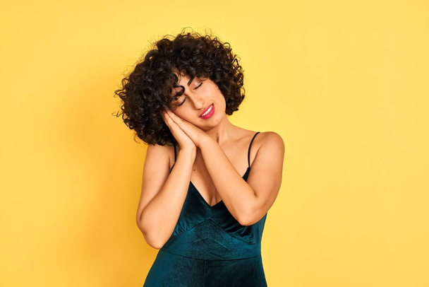Young arab woman with curly hair wearing elegant dress over isolated yellow background sleeping tired dreaming and posing with hands together while smiling with closed eyes. - Zdjęcie, obraz