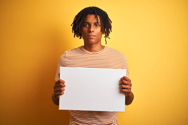 Afro amerykański człowiek z dredy trzyma banner na białym tle żółtym z pewnym wyrazem na inteligentne myślenie twarzy poważne - Zdjęcie, obraz