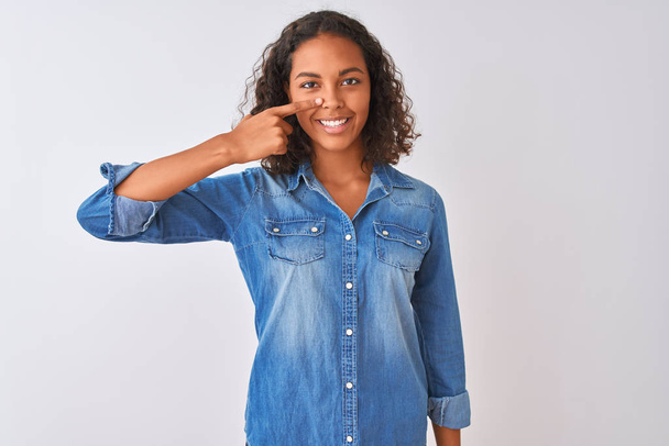 Young brazilian woman wearing denim shirt standing over isolated white background Pointing with hand finger to face and nose, smiling cheerful. Beauty concept - Photo, image