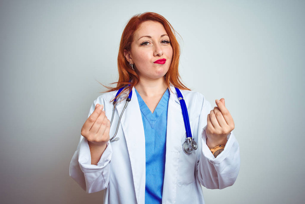 junge rothaarige Ärztin mit Stethoskop über weißem isoliertem Hintergrund macht Geldgeste mit den Händen, verlangt Gehaltszahlung, Millionärsgeschäft - Foto, Bild